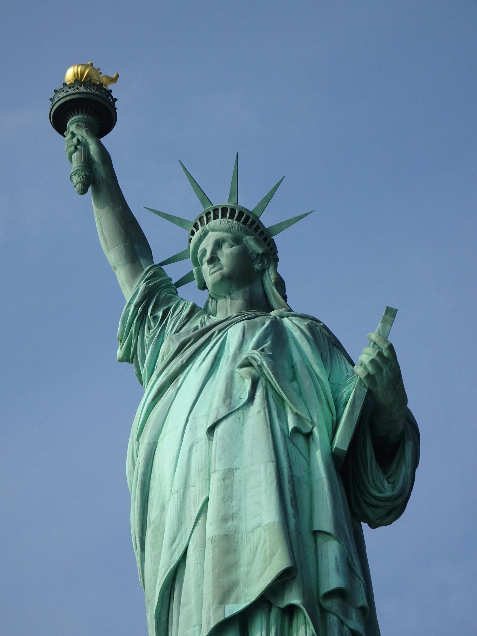 "Photographie de la Statue de la Liberté se dressant fièrement sur Liberty Island sous un ciel bleu clair."